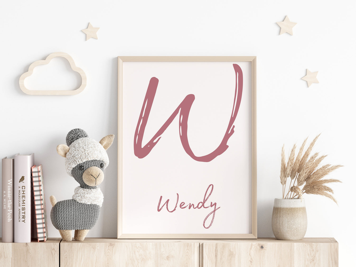 A personalized nursery wall art print featuring a large handwritten-style initial and cursive text below, framed and placed on a wooden shelf. The decor includes a plush llama toy, books, a vase with dried grass, and wooden star and cloud accents.