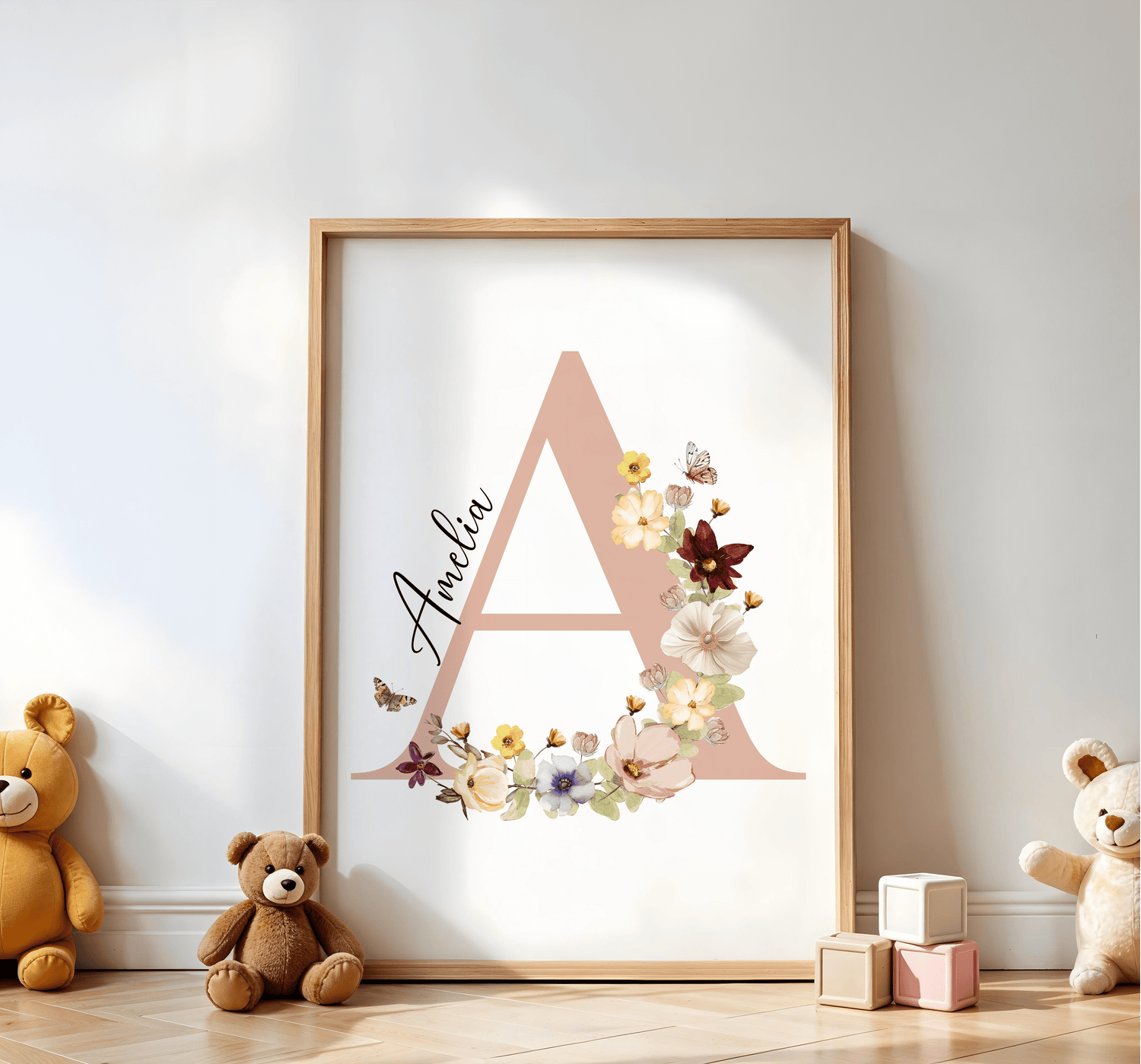 A framed nursery wall art print featuring a large pastel initial adorned with delicate flowers and butterflies, paired with elegant cursive text. The setup includes plush teddy bears and wooden blocks, creating a warm and playful atmosphere.