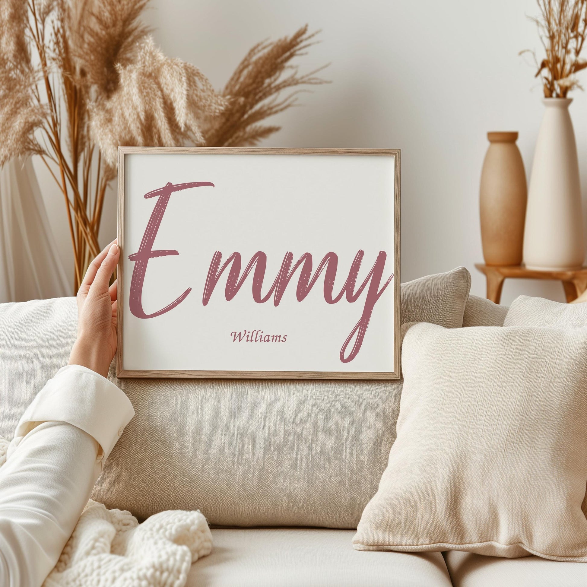 Framed custom baby name sign with the name Emmy Williams in pink brush font, held by a hand against a cozy, neutral-colored background with dried flowers.