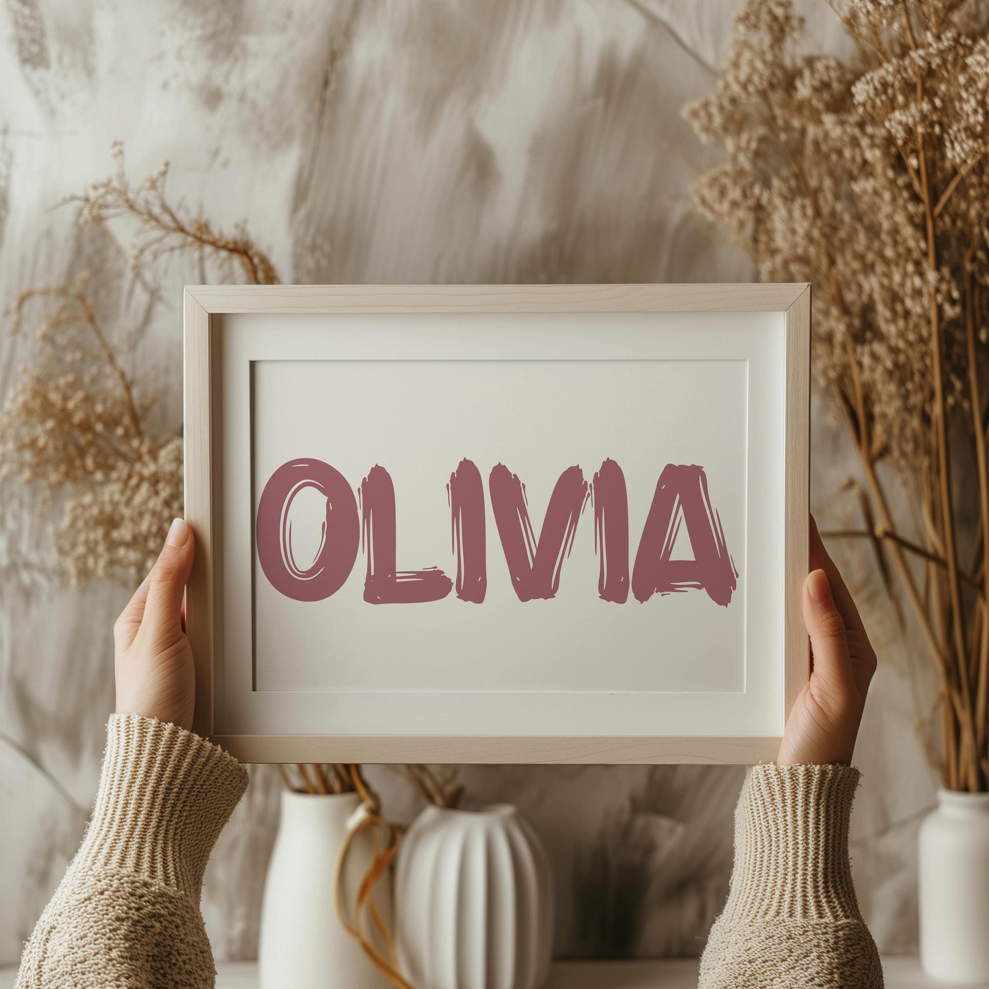 Framed personalized baby name sign with the name Olivia in bold pink brush font, held by hands against a background with neutral decor elements.