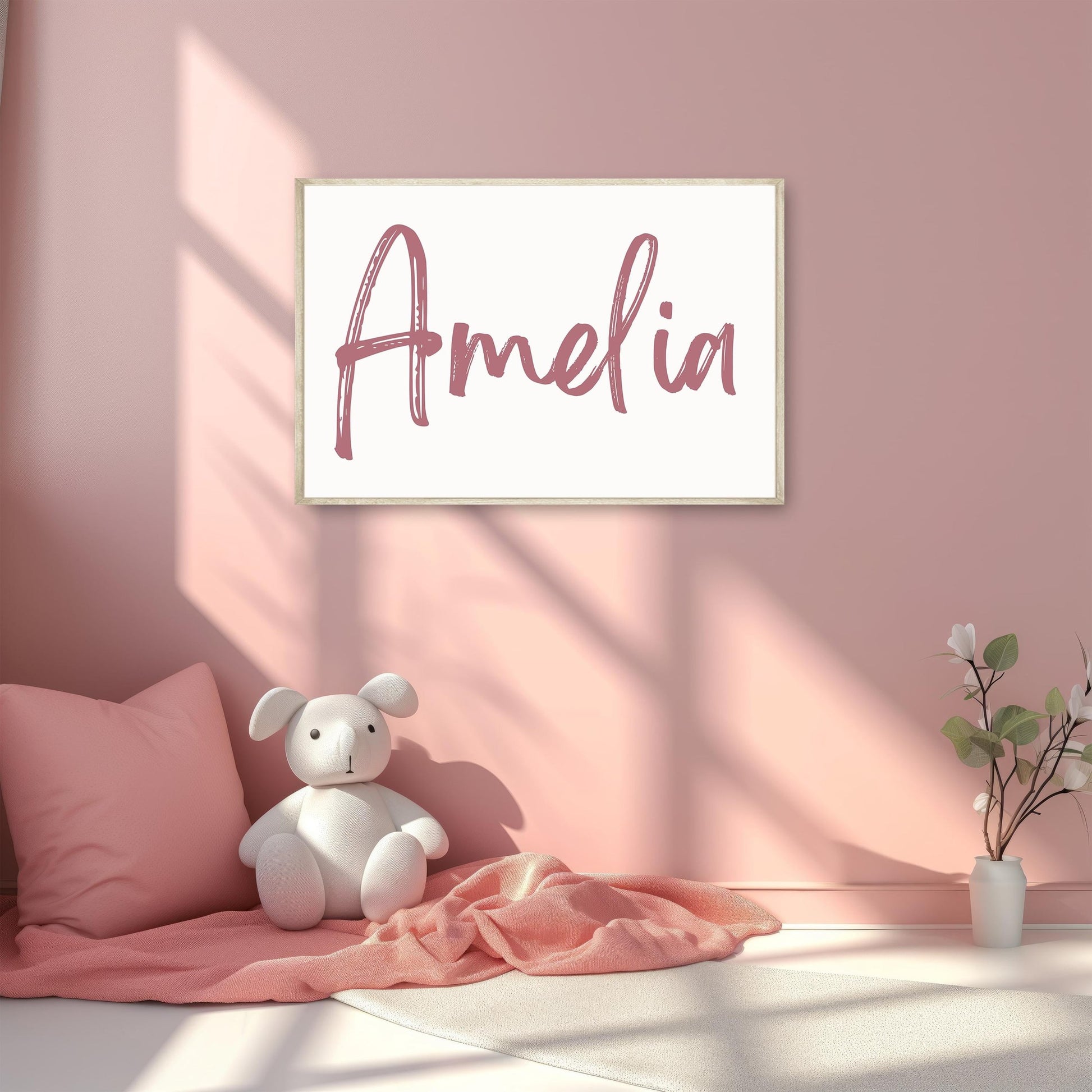 Nursery wall art featuring the name Amelia in pink brush font, framed and displayed in a cozy, pastel nursery setup with a teddy bear and blanket.